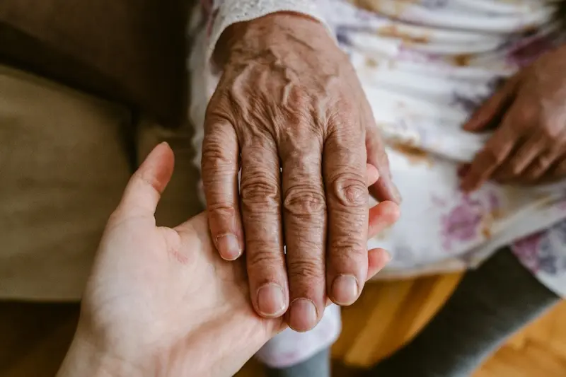 A close-up, top-down view of a younger person's hand gently cupping and supporting the weathered, wrinkled hand of an elderly person.