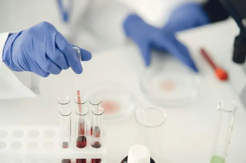A laboratory worker wearing blue gloves uses a pipette to handle red liquid in a rack of test tubes.