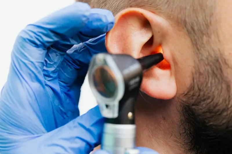 A close-up of a medical professional wearing blue gloves using an illuminated otoscope to examine a patient's ear canal.
